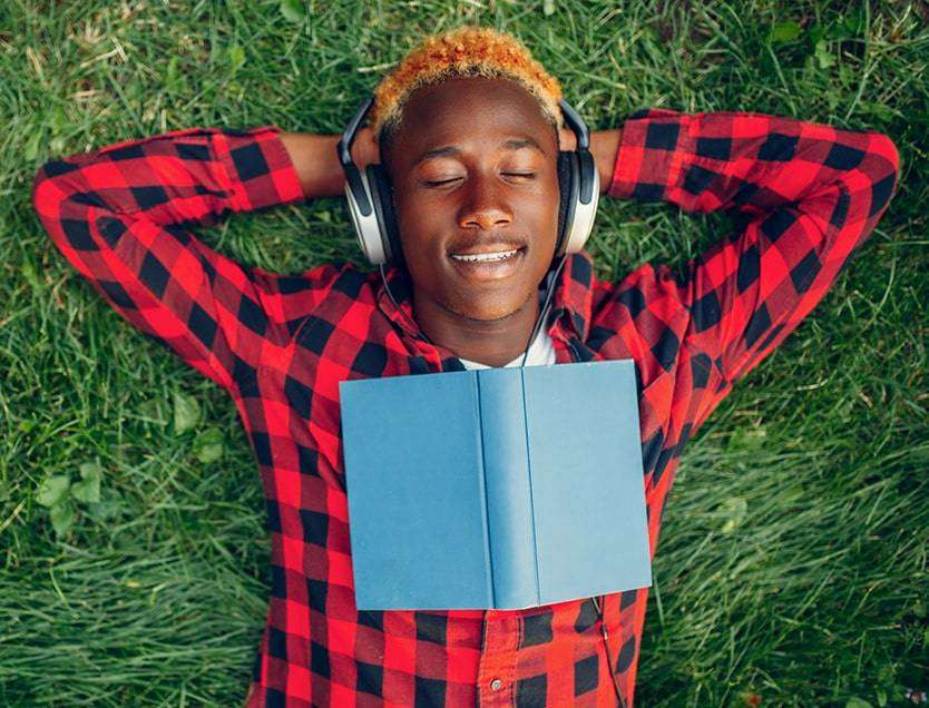 Man of colour with dyed ginger hair lying on his back on some grass with headphones on, his eyes closed and a book across his chest.
