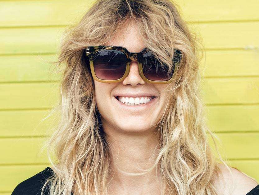 Headshot of white woman with blonde curly hair, wearing sunglasses and smiling against a yellow wooden hut.