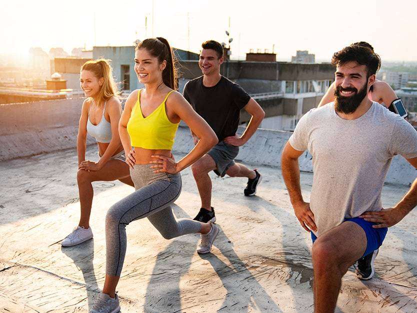 Group of 5 white people, all in workout clothes and smiling, in a lunge position on the roof of a building in the sunshine, overlooking a city.